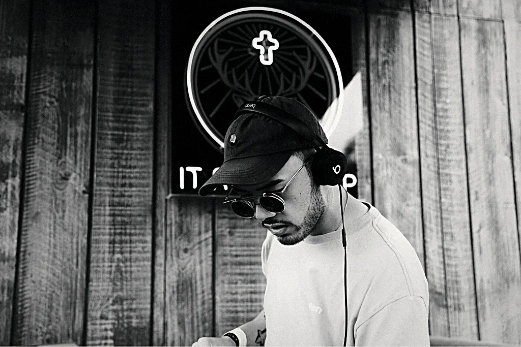 BDJ at the decks in black and white, head down beneath a circular neon sign