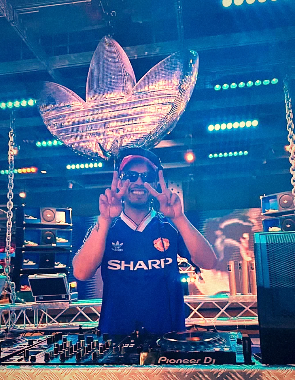 BDJ on a festival stage under rig lighting, arms raised behind the decks
